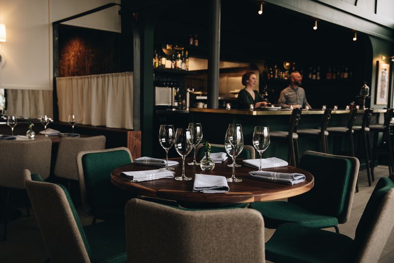 A round wood table with green chairs around it and wine glasses and napkins on top of it, with a bar and other tables visible in the background.