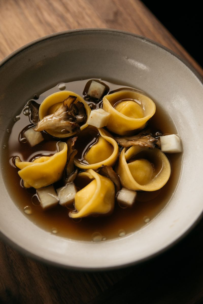 Yellow tortellini floating in brown acorn broth in a beige clay bowl on a wooden table. 