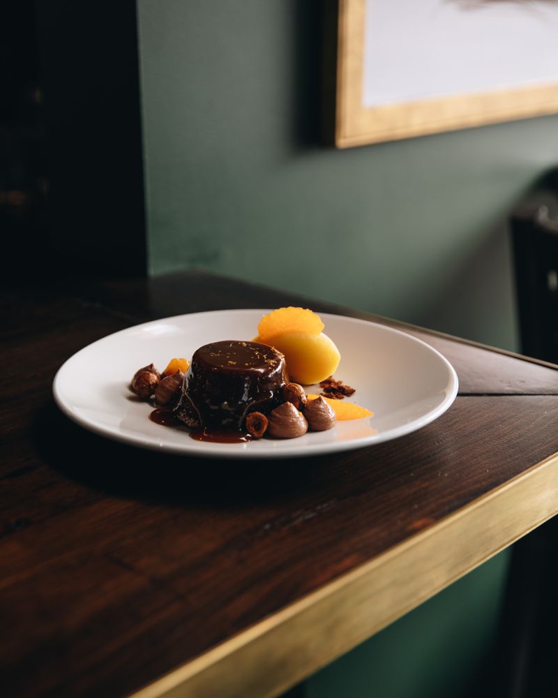 A white plate on a dark wood counter with a small cake covered in dark toffee sauce, with a quenelle of orange sorbet on the side. 