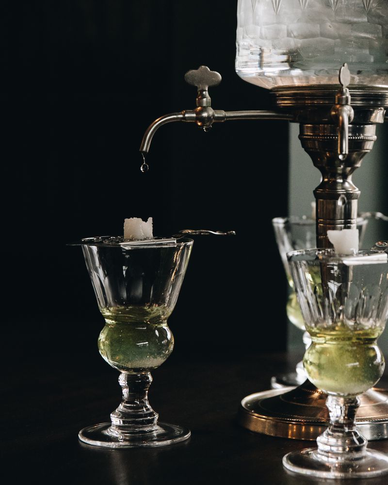 A glass with a small amount of green liquid at the bottom and a strainer with a sugar cube on top of it. A glass pitcher is dripping water onto the sugar cube. 