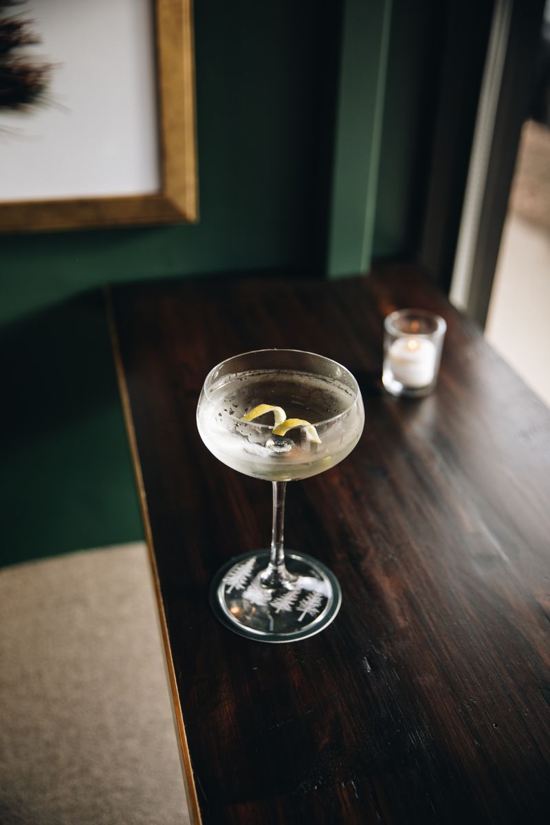 A tall coupe glass filled with a clear cocktail on a dark wood counter, in front of a deep green wall. 