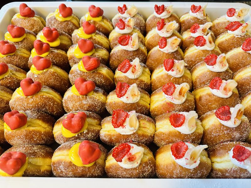 Rows of brioche doughnuts, all topped with dollops of cream and either a halved raspberry or a red heart, in a white bin. 