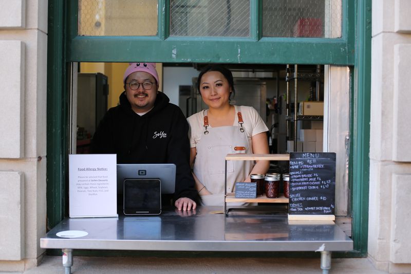 A person wearing glasses, a black sweatshirt, and a pink beanie, and another person wearing a white T-shirt and a beige aprom, standing at a counter through a green-painted window. 