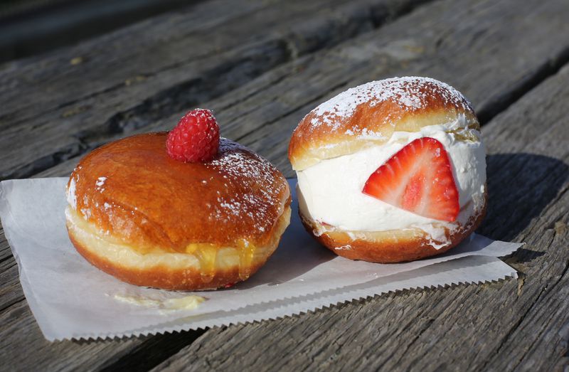 Two doughnuts, one with a hard sugar glaze and a raspberry on top; one split in half and filled with cream with a slice strawberry pressed into the cream, sitting on a white paper on a wooden table. 