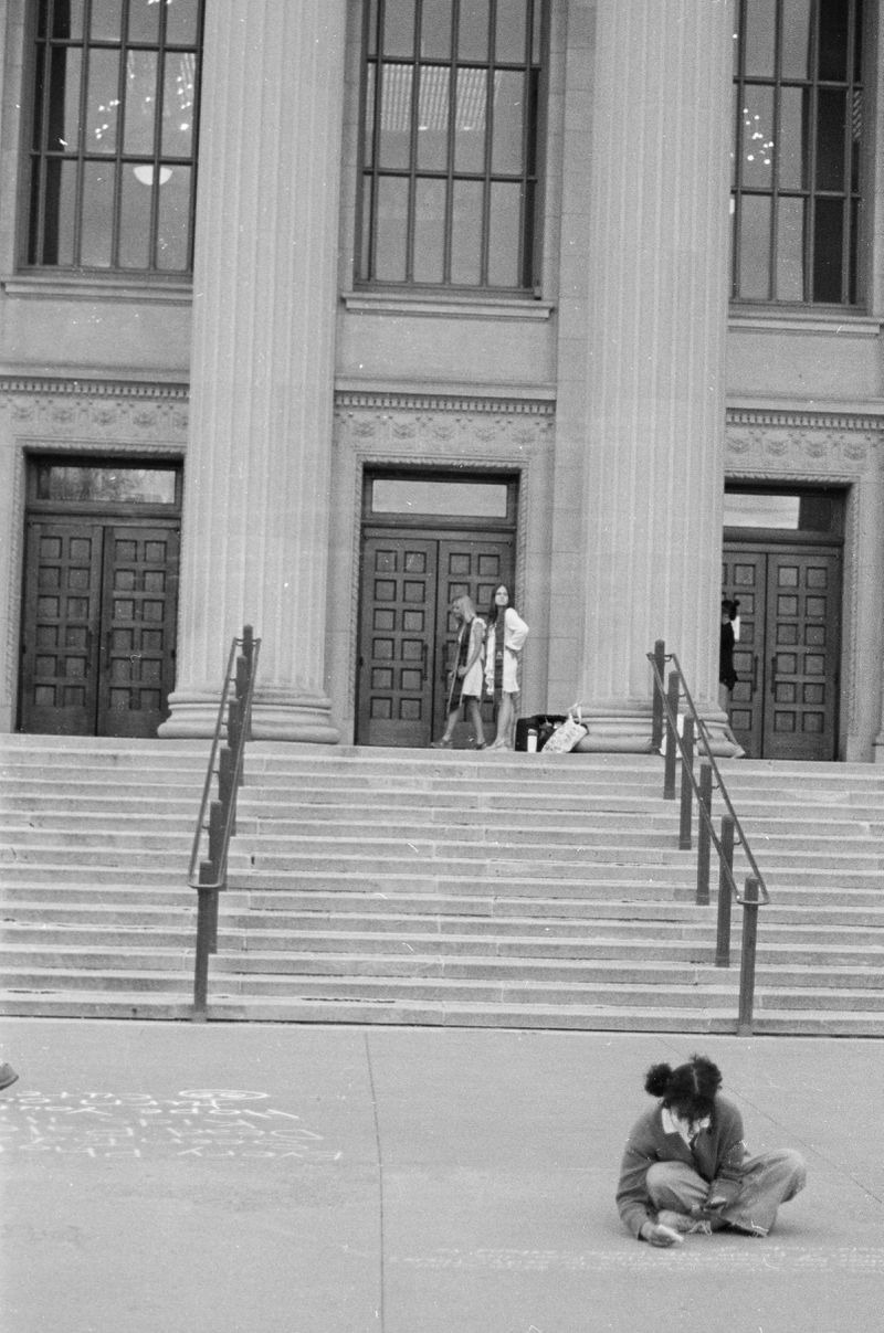 Two women in graduation regalia (white graduation gown and darker colored stoles) are pictured standing at the top of the steps of Northrup Hall. In the foreground, a young girl with two side-buns writes messages on the sidewalk below.
