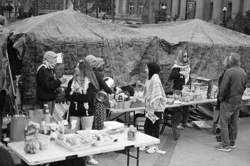 A group of people, some wearing keffiyehs around their heads, milling about in an area with a number of long white tables laden with food, drinks, and snacks, in front of a long canvas tent. 