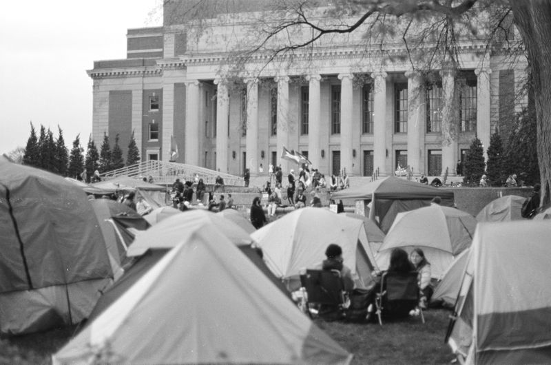A group of camping tents foreground a large university building with 10 identical pillars at its entrance. The collection of tents, some with students sitting near or in them, formed a since-disbanded encampment at the University of Minnesota.