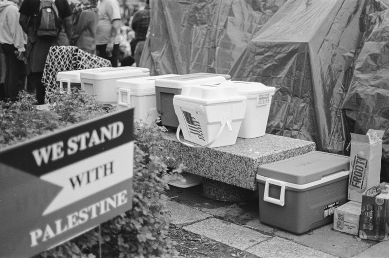 A row of coolers, one marked with an American flag, sitting on a marble bench. In the foreground, there’s a sign with the Palestinian flag that reads “We Stand With Palestine.”