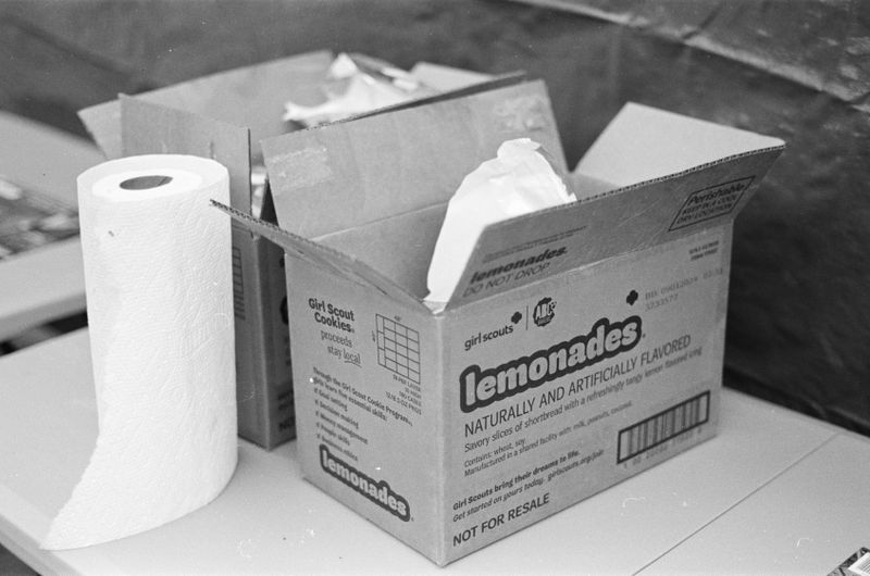 A cardboard box of cookies reading “Lemonades”sitting on a plastic table next to a roll of paper towels and a similar cardboard box directly behind it. 