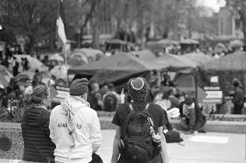 Three people sitting with their backs to the camera, one with a keffiyeh around their shoulders, another wearing a yarmulke, looking out at a collection of tents and people gathered at the encampment. 