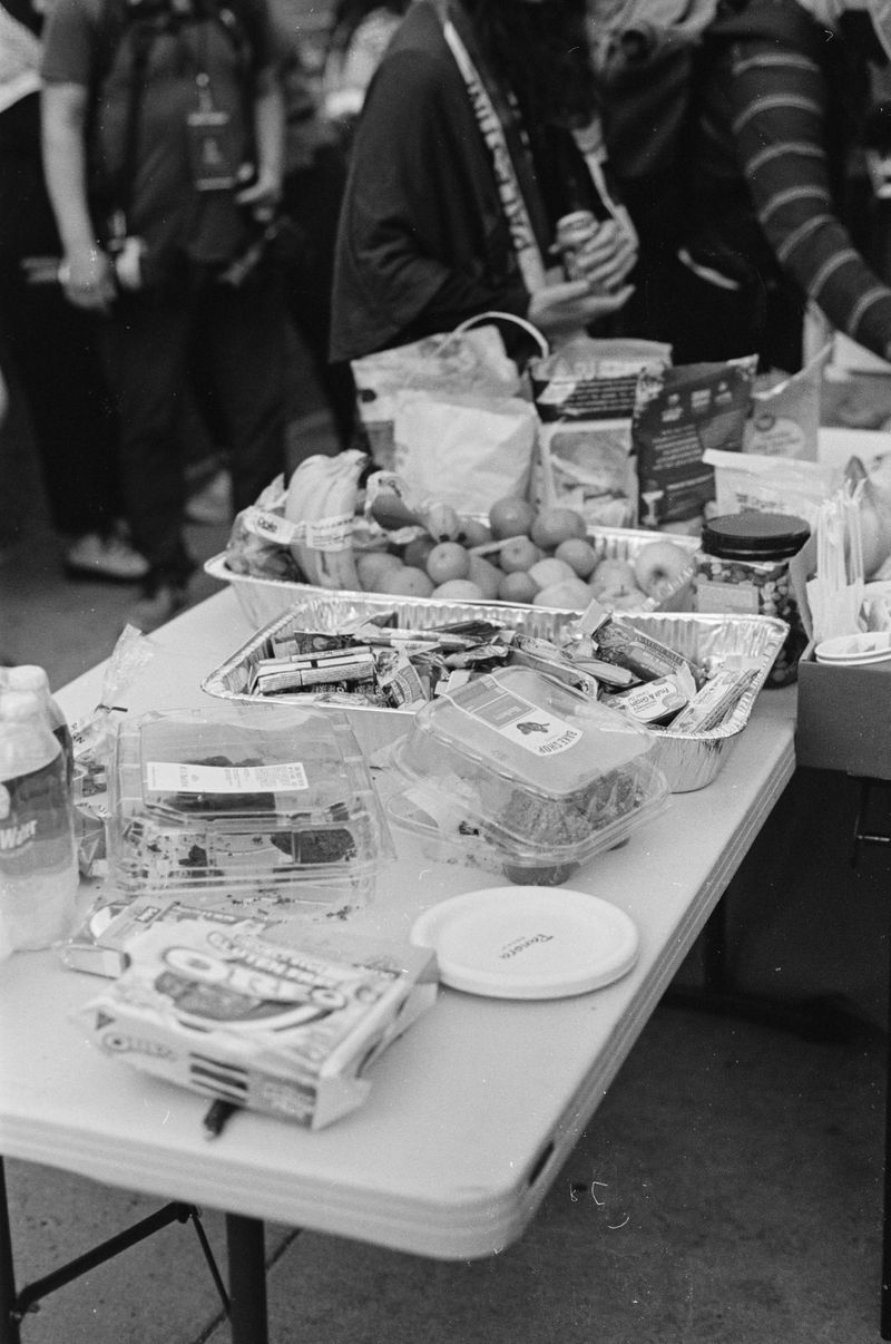 A white folding table with tin catering trays holding fruits (bananas, apples) and protein bars, as well as clear plastic trays of muffins and other baked goods.