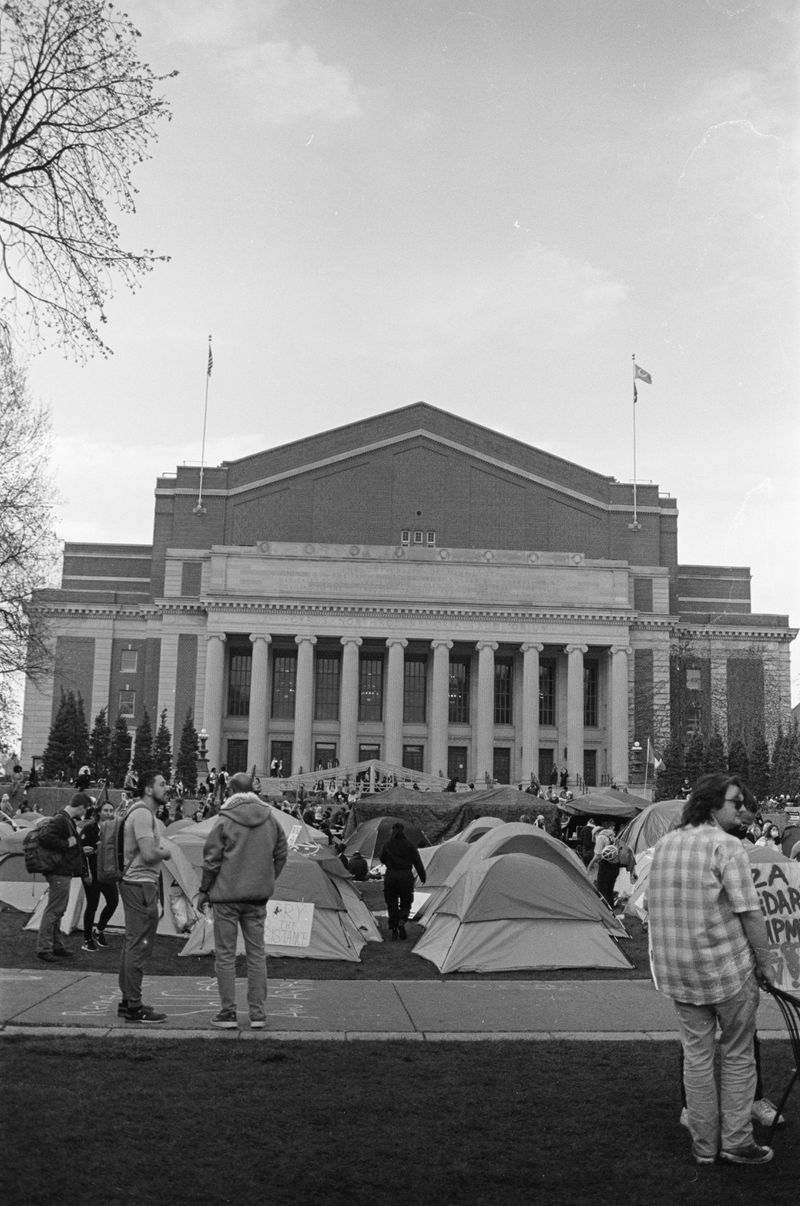 The exterior of Northrop Hall, a tall stone building with tall pillars in front, and an assortment of tents and people standing on the lawn in front, in the foreground. 