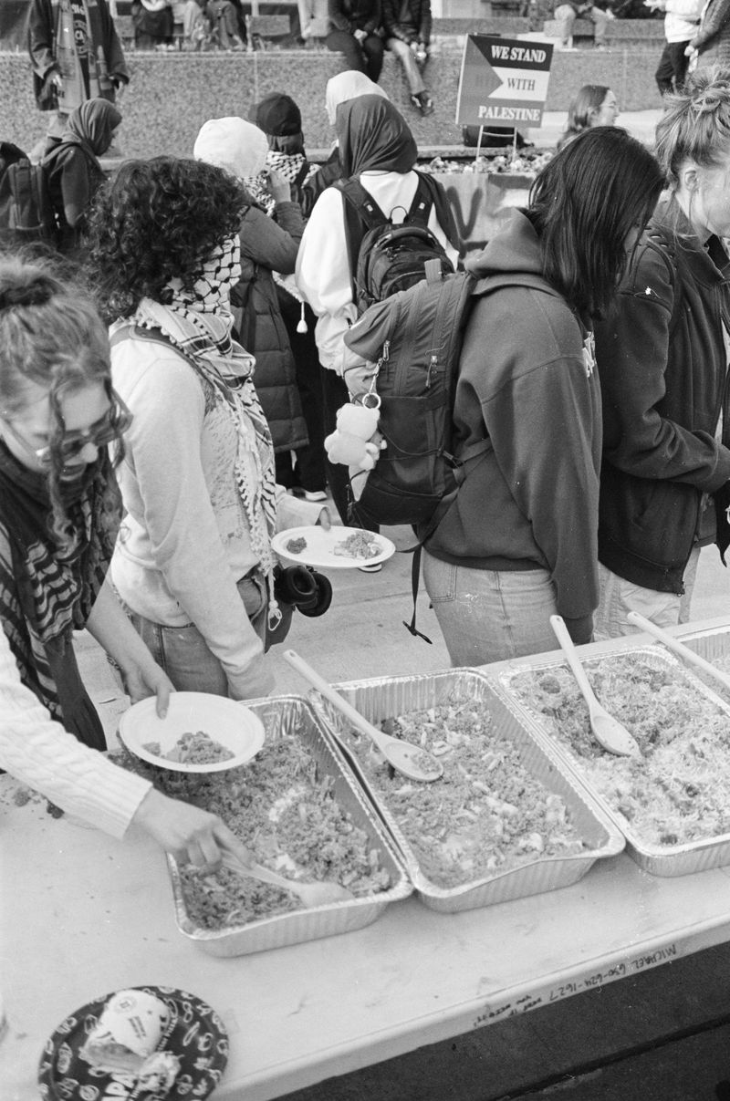 Students wearing sweatshirts, backpacks, and keffiyehs standing in line at a long table set with silver trays of biryani and fried rice, holding plates and scooping food onto them. 