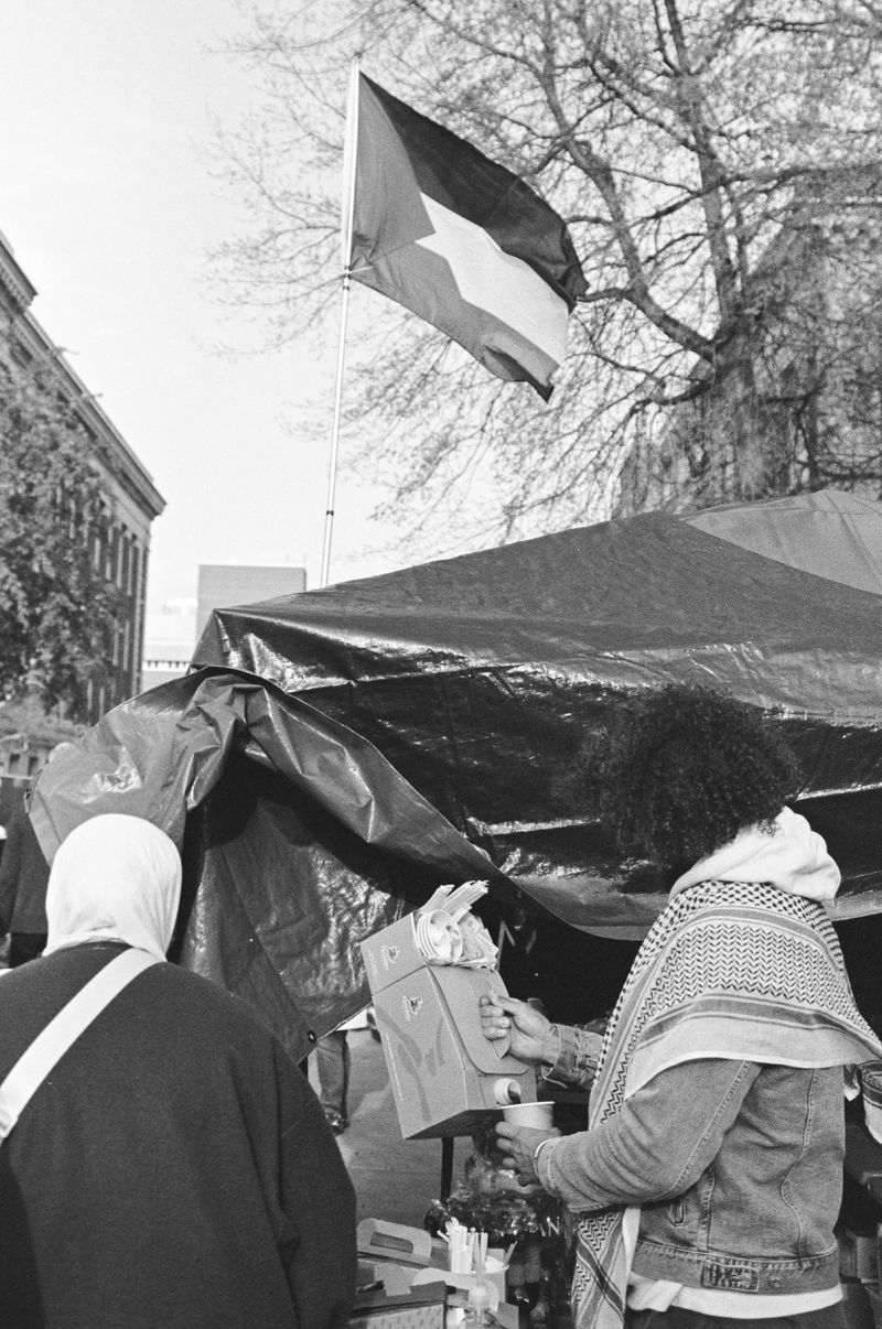 Two people with their backs to the camera standing outside a tarp tent, one, with a keffiyeh wrapped around their shoulders, holding a cardboard container of coffee and pouring some into a cup. 