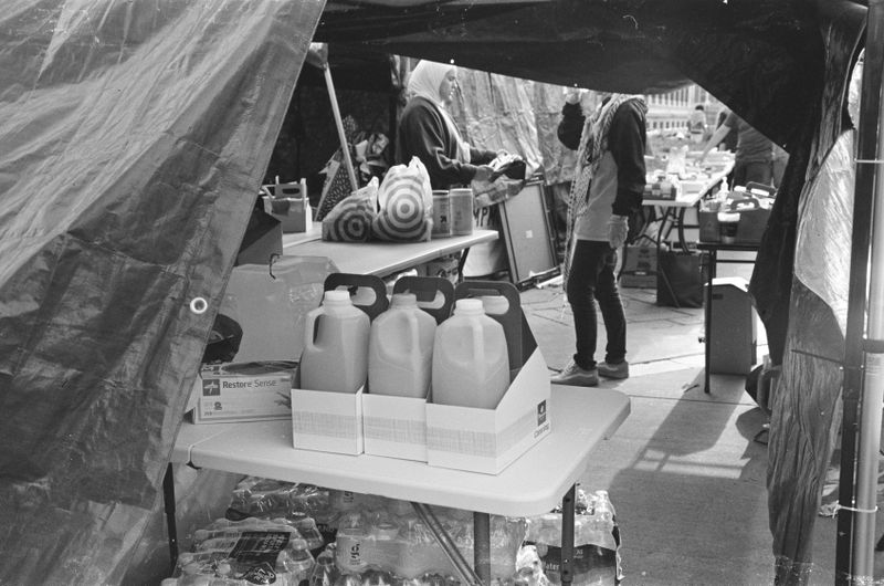 Half-gallon jugs of juice sitting in cardboard boxes on a white table inside a makeshift tent with other bags of supplies, and a person in an apron half-visible in the background. 