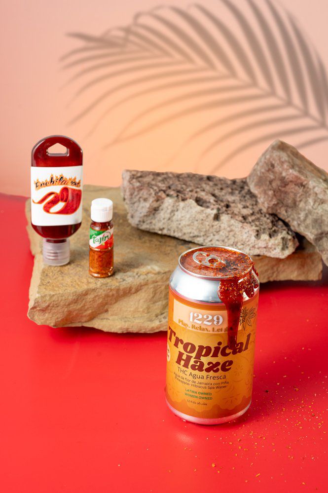An orange can with the words “Tropical Haze” and bright red chamoy dripping from the top of the can. Behind it there are three flat rocks stacked on top of each other; there’s a small bottle of tajin and a squeeze bottle of chamoy. All of this sits on a bright red surface; the background is light pink with the outline of a palm frond. 