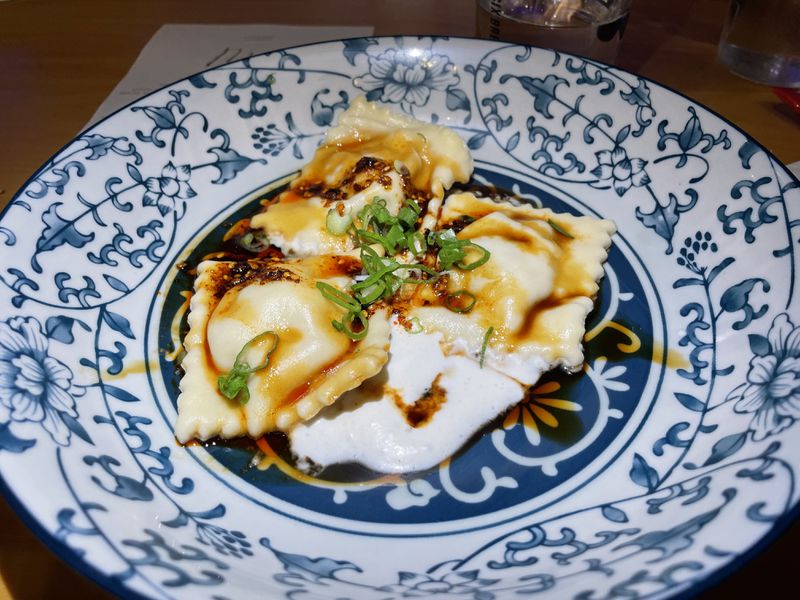 Four ravioli sitting in a drizzle of chili soy sauce over a pool of white mushroom cream on an intricately patterned blue and white plate. 