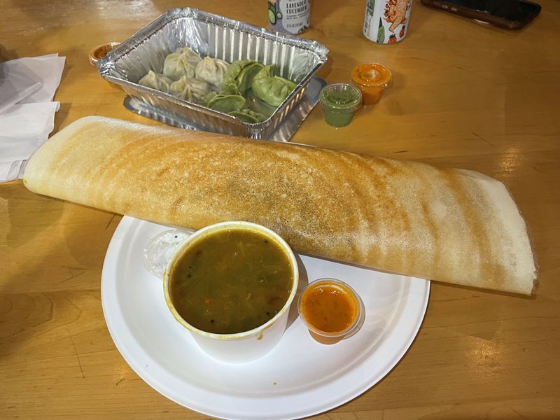 A large dosa folded and sitting on a white paper plate next to a small dish of sambar, with a foil dish of moos in the background on a wooden table.
