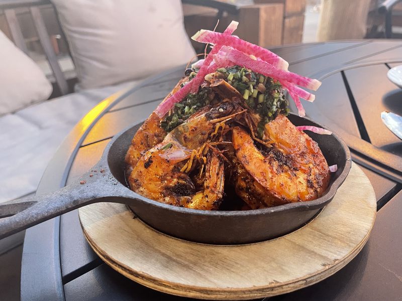 A small cast-iron skillet of grilled prawns topped with chimichurri and sliced watermelon radish sitting on a black table. 