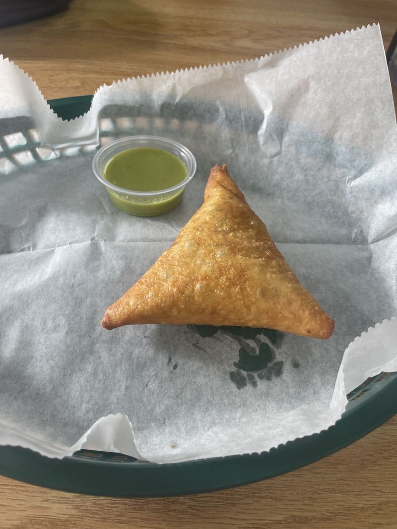 A single triangular sambusa sitting in a green plastic basket lined with white paper and a small plastic dish of bright green sauce. 