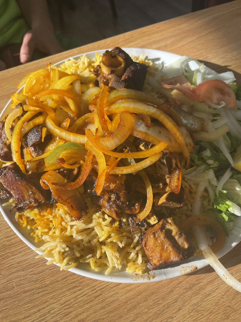 A round white plate heaped with rice and morsels of goat meat under sauteed peppers and onions with a side salad, sitting in sunlight on a wooden table. 