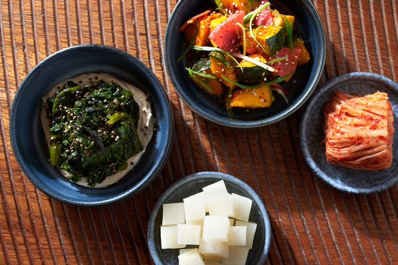 Four small blue dishes set on a wooden table, each holding a different banchan: One of mustard greens over a creamy tahini sauce; one a dongchimi radish kimchi, one a bright red cabbage kimchi, and one of kabocha squash and radish slices dotted with sesame seeds and topped with scallions. 