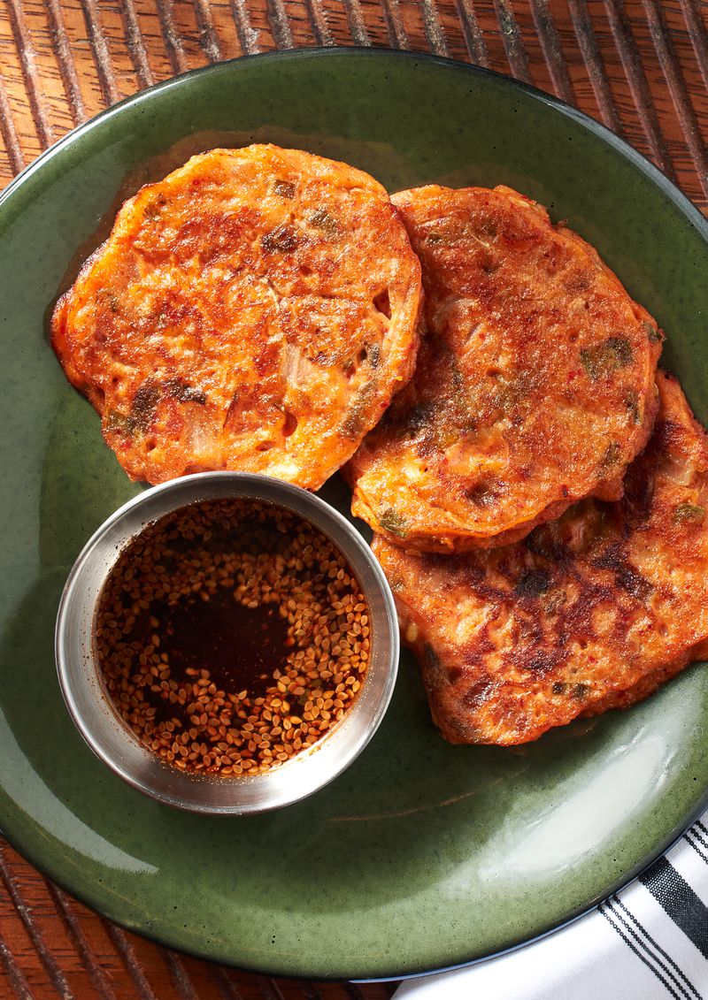 Three bindaetteok, which are Korean pan-fried pancakes made from mung beans, rice, and kimchi, resting on a circular green plate with a small metal dish of soy sauce with sesame seeds floating in it, on a wooden table with grooves in it. 