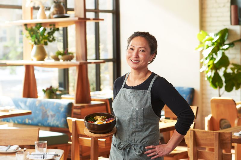 Chef Ann Kim wearing a grey apron with a black long-sleeve shirt, holding a stone bowl bibimbap in one hand with the other hand on her him, smiling at the camera. In the background is the interior of her restaurant, Kim’s, with large sunny windows and wooden tables and chairs. 