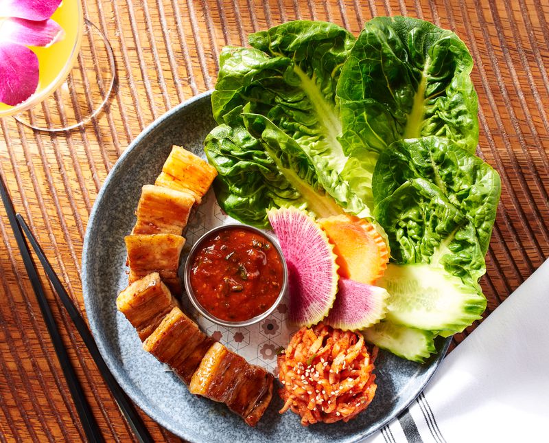 A marled grey plate holding slices of pork belly, radishes and cucumbers, kimchi, a dish of ssamjang, and large lettuce leaves on a wooden table with curved grooves in it. 