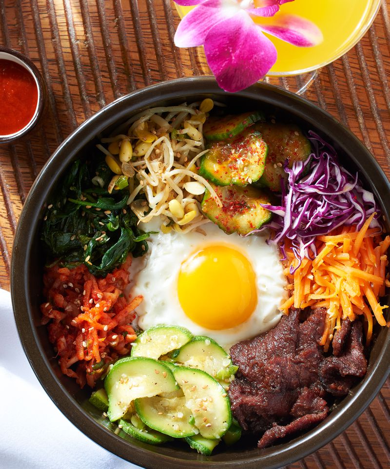 A large dark grey round bowl with a sunny-side-up egg at the center and a circular arrangement of beef bulgogi and banchan like carrots, cabbage, and zucchini sitting on a wooden table, with the lip of an orange cocktail glass sitting beside it. 
