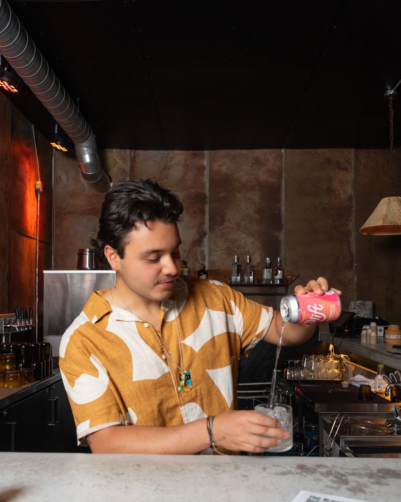 A person wearing a white and orange patterned shirt pouring a pink and orange can of hemp-infused sparking water in to a glass and standing behind a bar. 