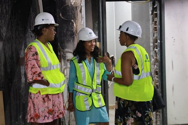 Three women in hardhats and neon vests chat at a construction site