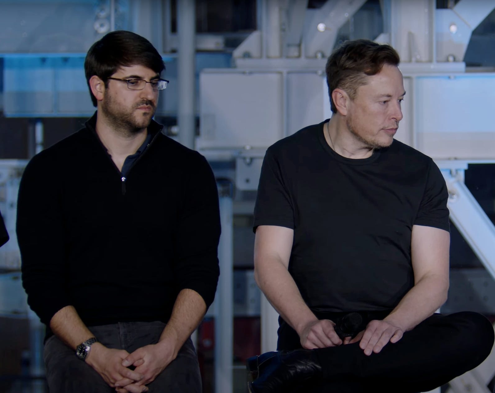 Zachary Kirkhorn wearing a black button-down shirt while sitting next to Elon Musk with his hands over his lap.