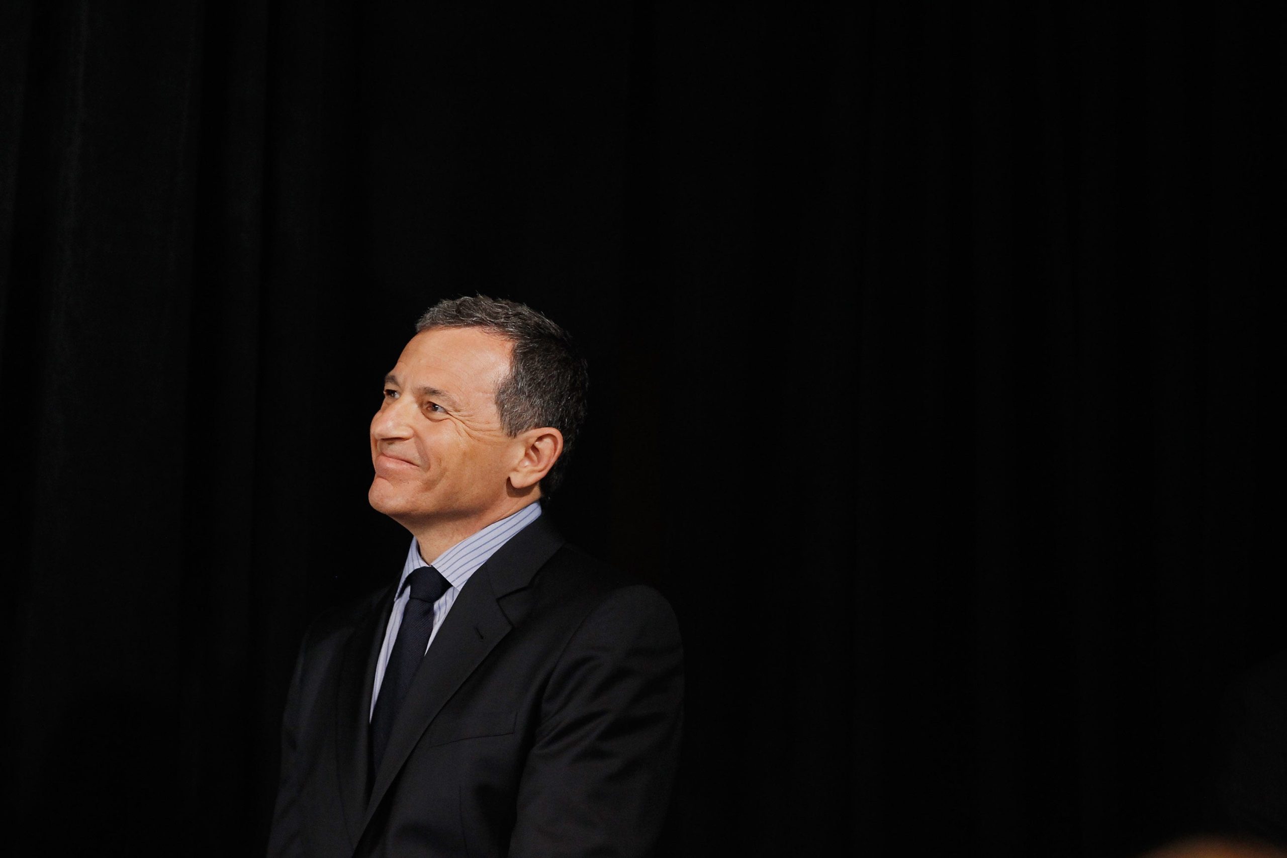 Bob Iger smiles off camera while wearing a suit in front of a black background.