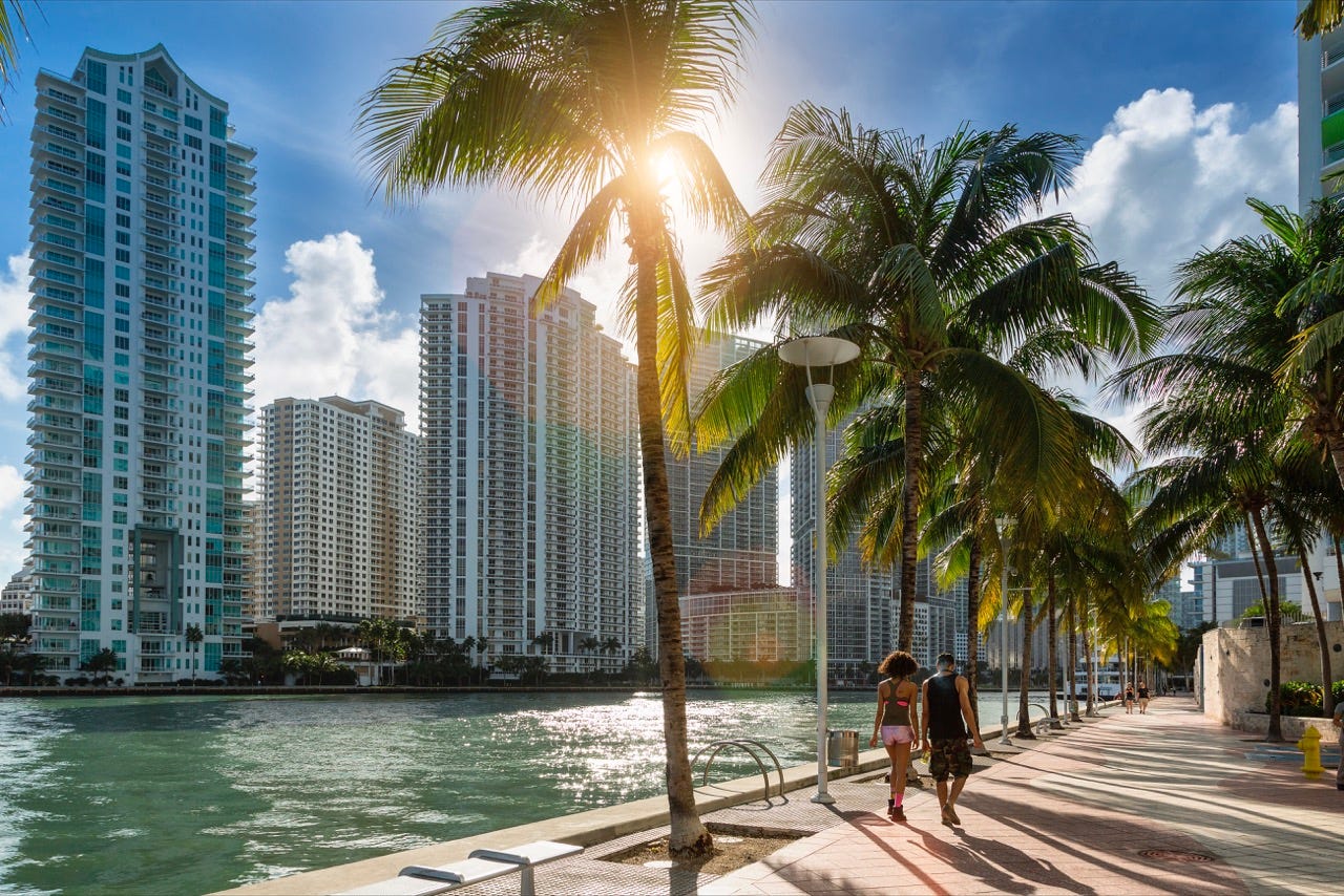 A couple walks down a palm-tree lined Miami boardwalk with towering white condos in the background.