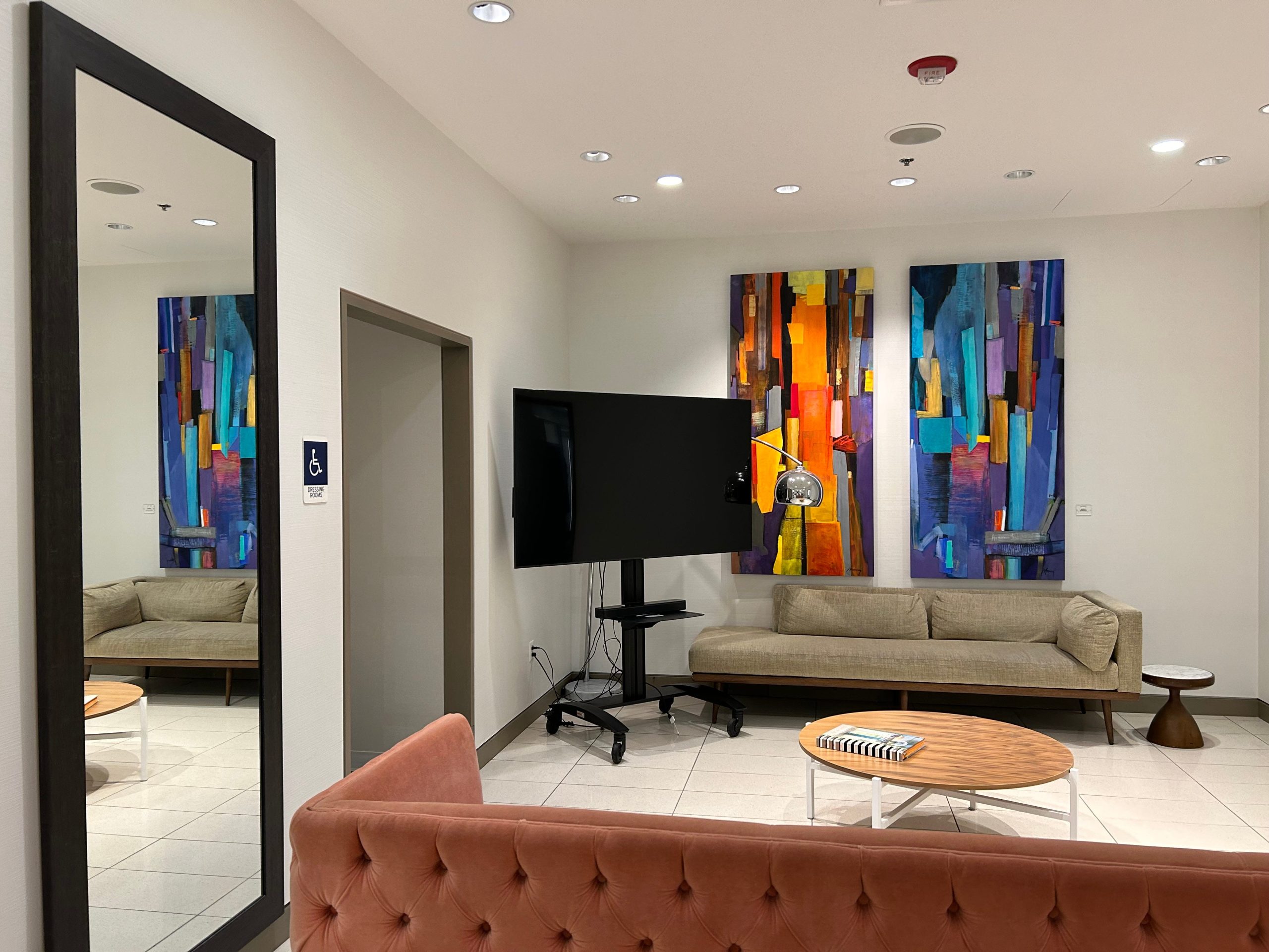 A waiting area outside a Nordstrom fitting room with two couches, a round table, a TV, and a mirror and paintings on the walls.