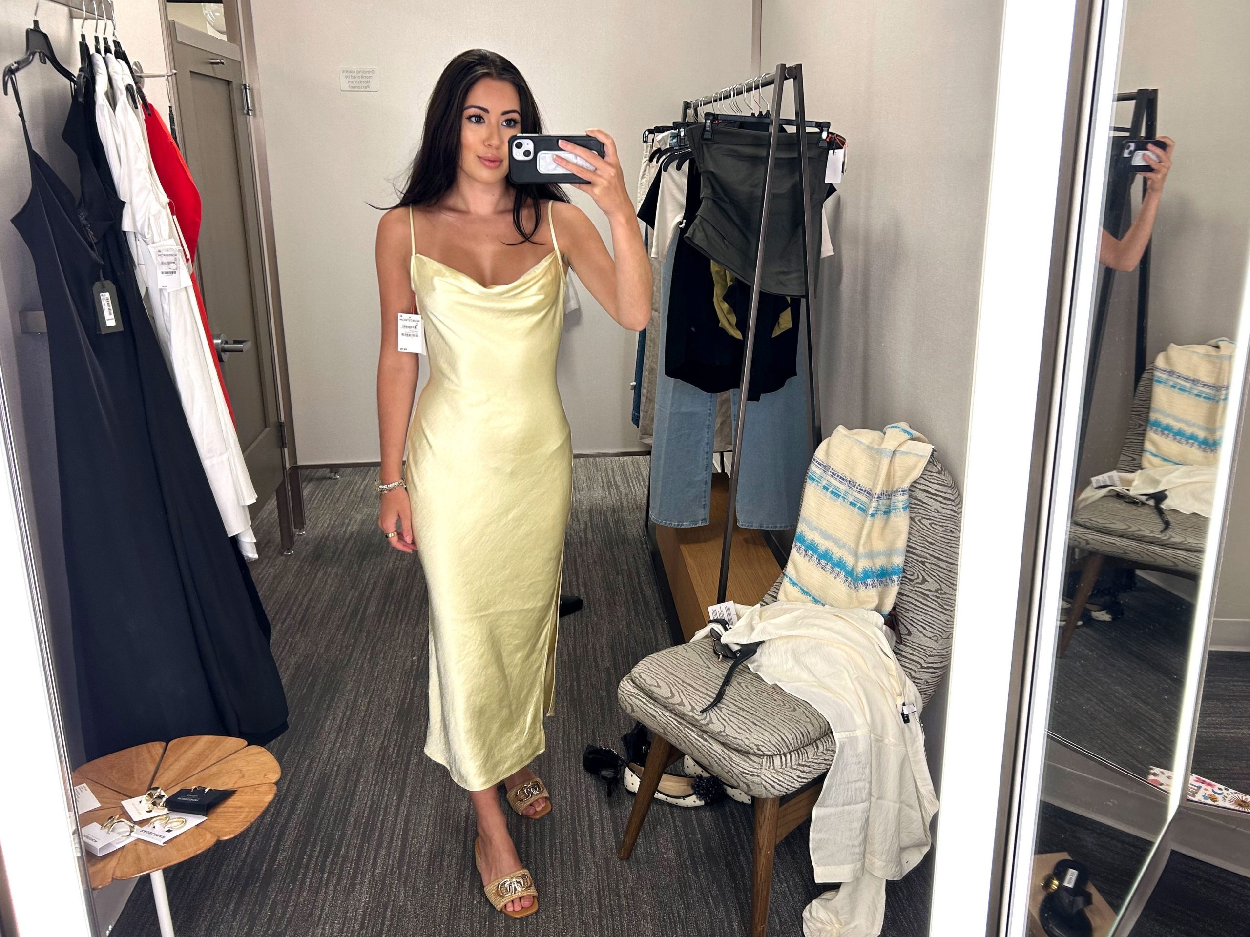 Lara wearing a yellow satin slip dress in a fitting room. She is taking a photo of herself with her phone, and clothes are scattered around her.