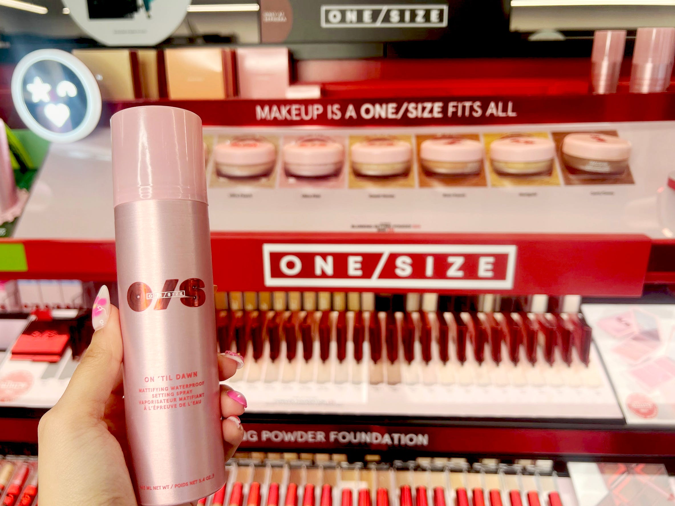 A hand with pink nails holds a large pink aerosol  can of setting spray with red One Size logo on it in front of One Size display at Sephora