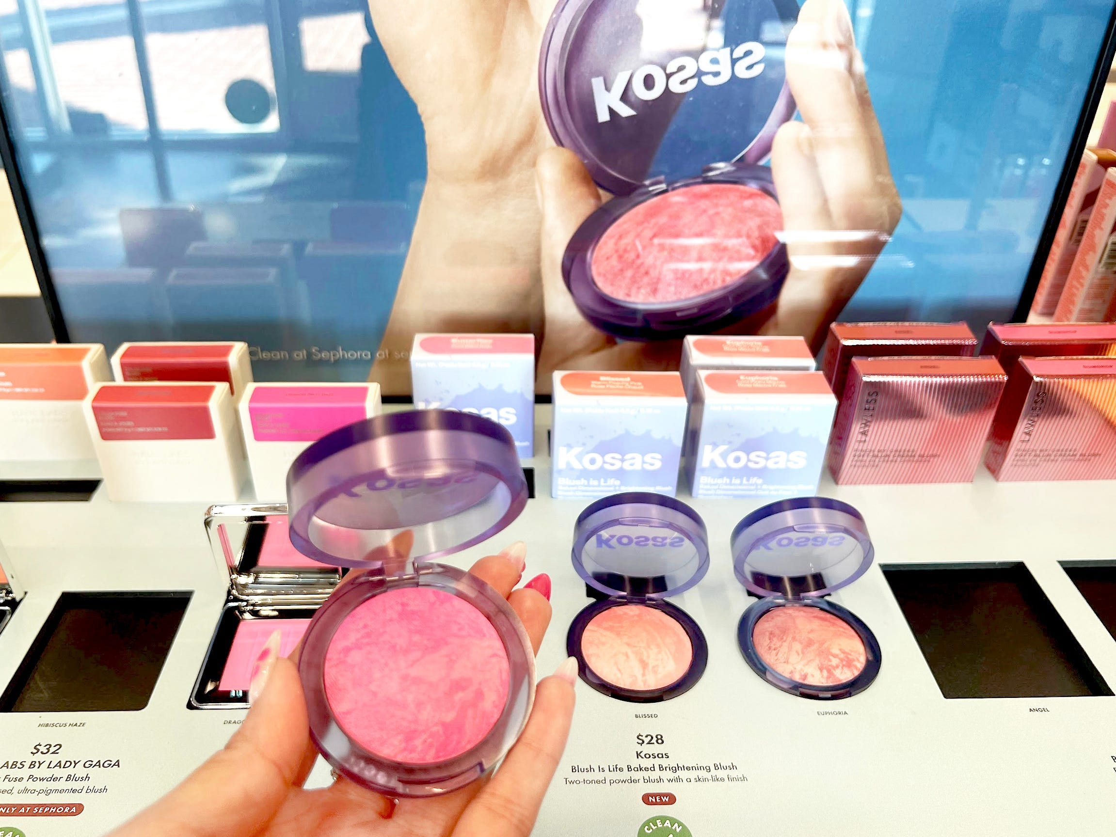 A hand holds a compact blush with bright-pink marbled press-powder product in front of a white display with Kosas blushes and a large image of Kosas blush