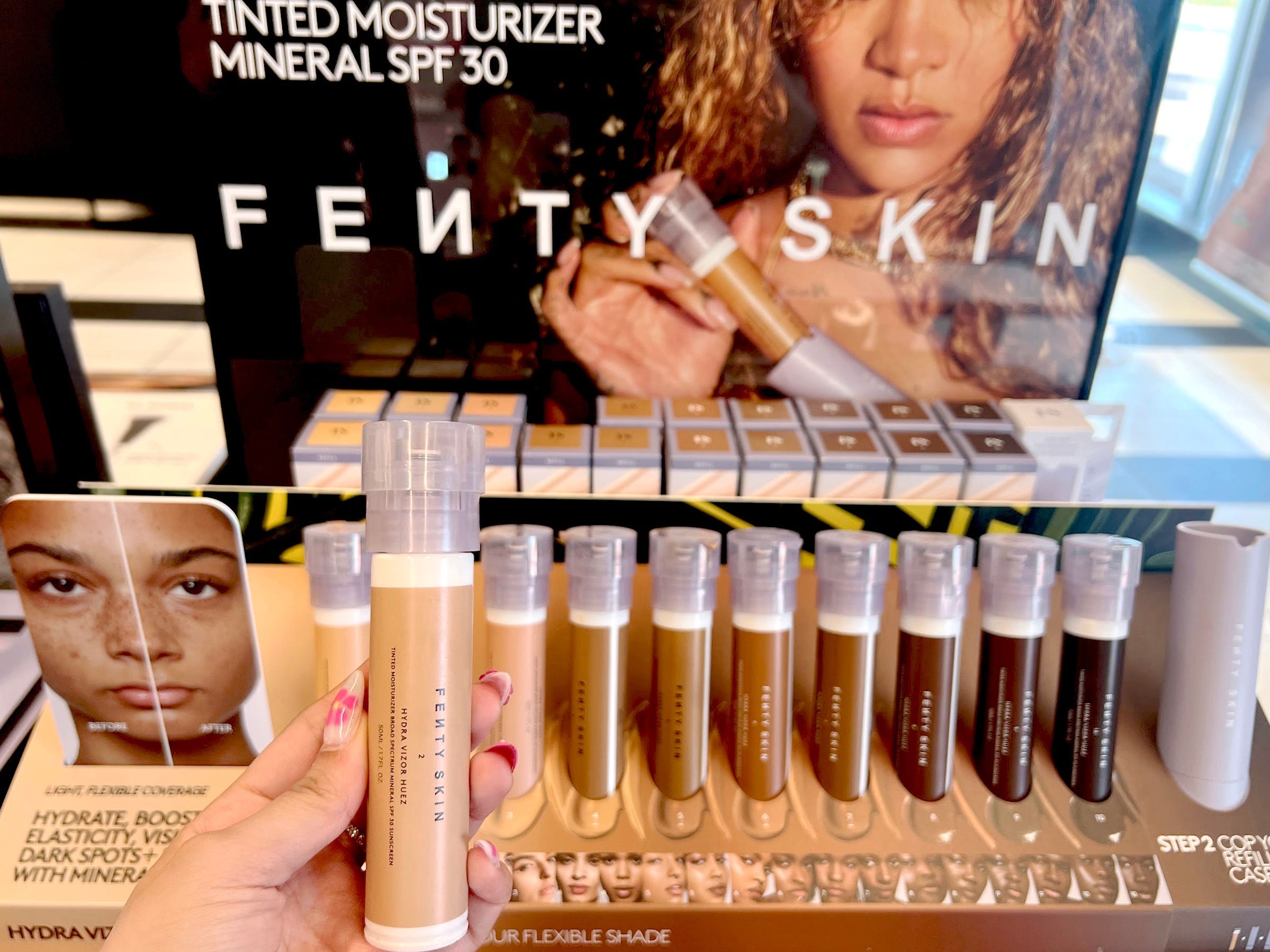 A hand holds a tan tube of Fenty Skin tinted moisturizer in front of a Fenty Skin display with several tubes with purple caps and a picture of Rihanna