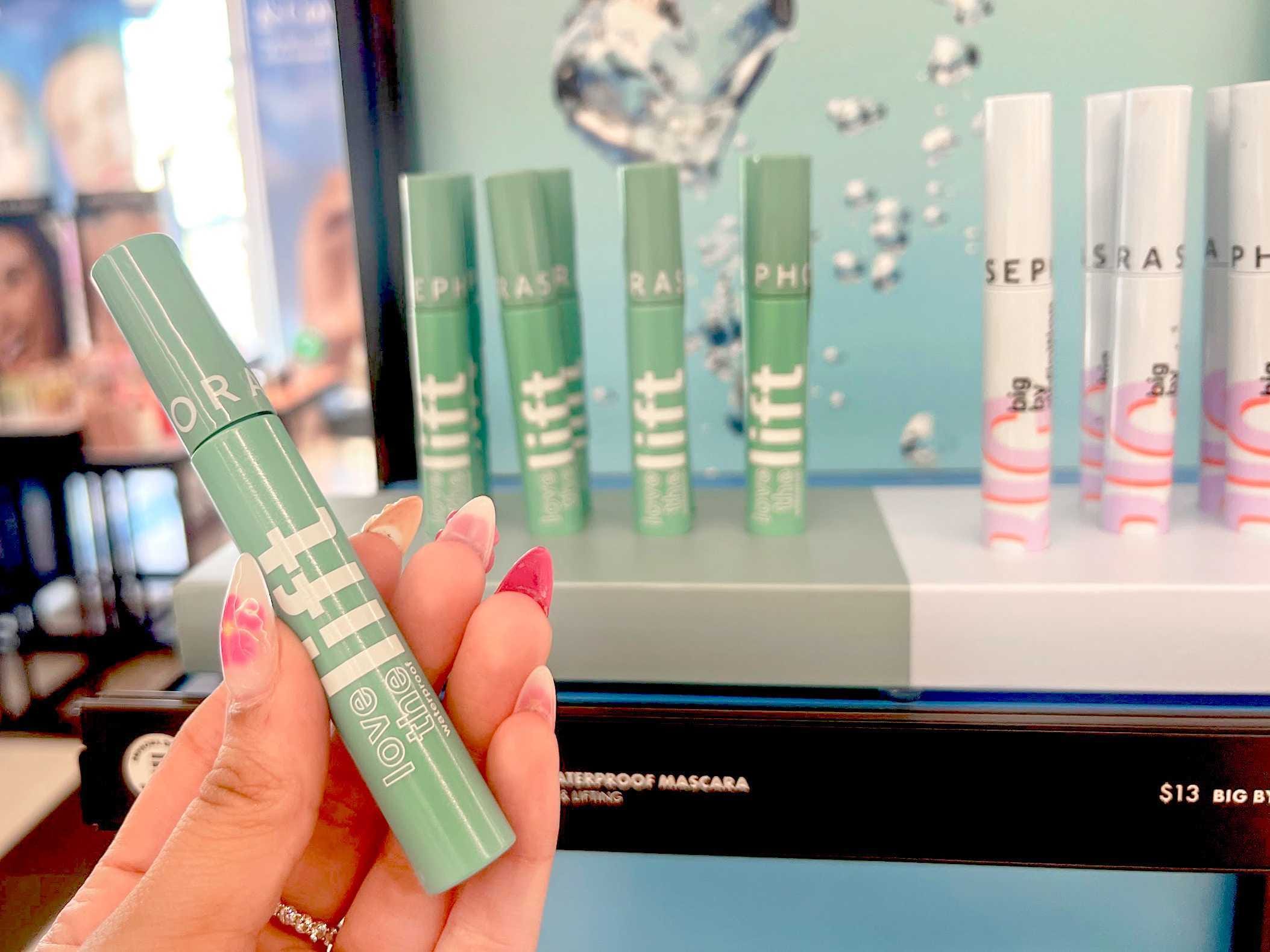 A hand holds a green tube of mascara with white lettering spelling out "Love the Lift" in front of a green and white mascara display