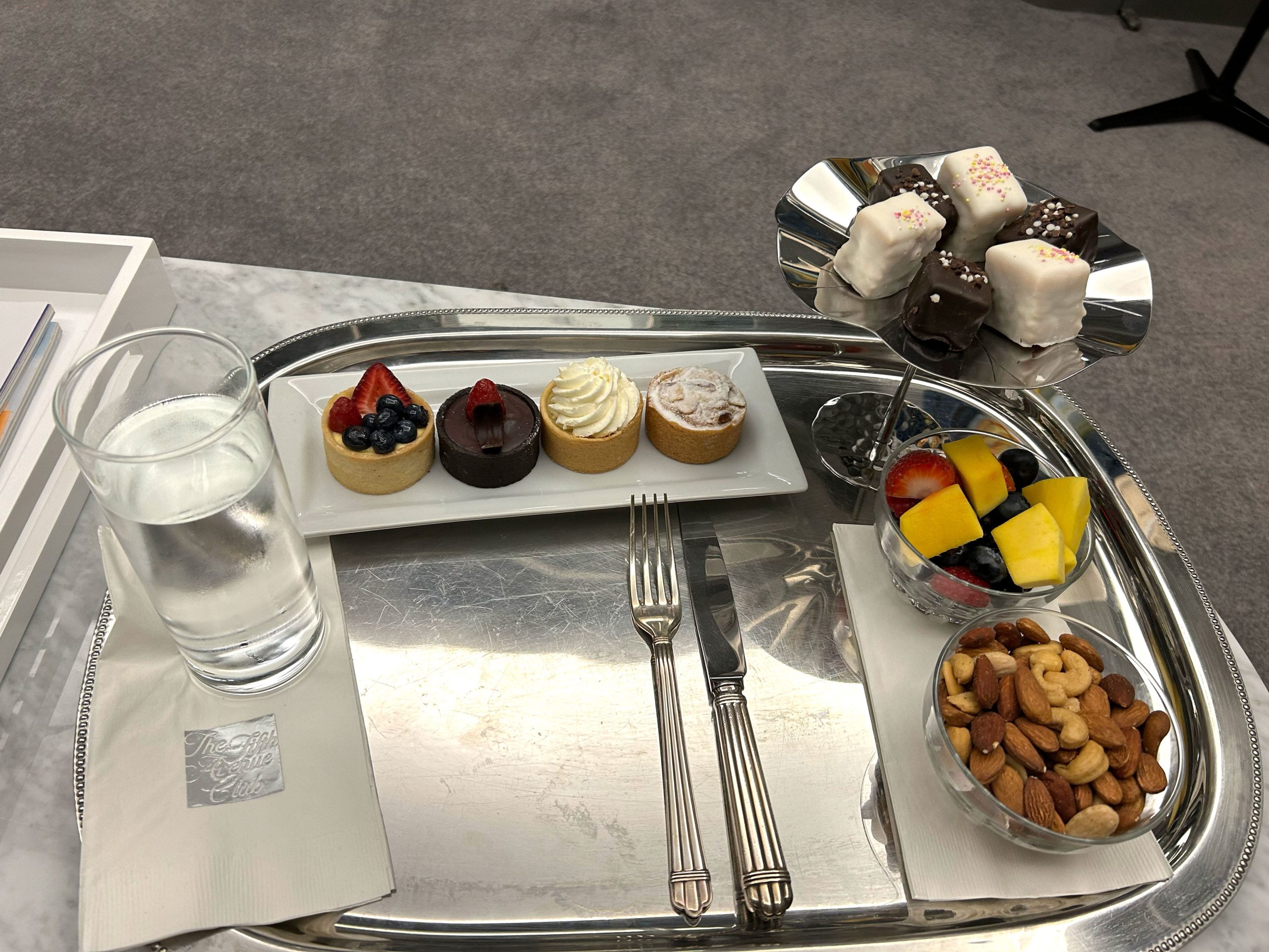 A silver platter with mini cakes, fruit, nuts, a glass of water, silverware, and a napkin that says "The Fifth Avenue Club."