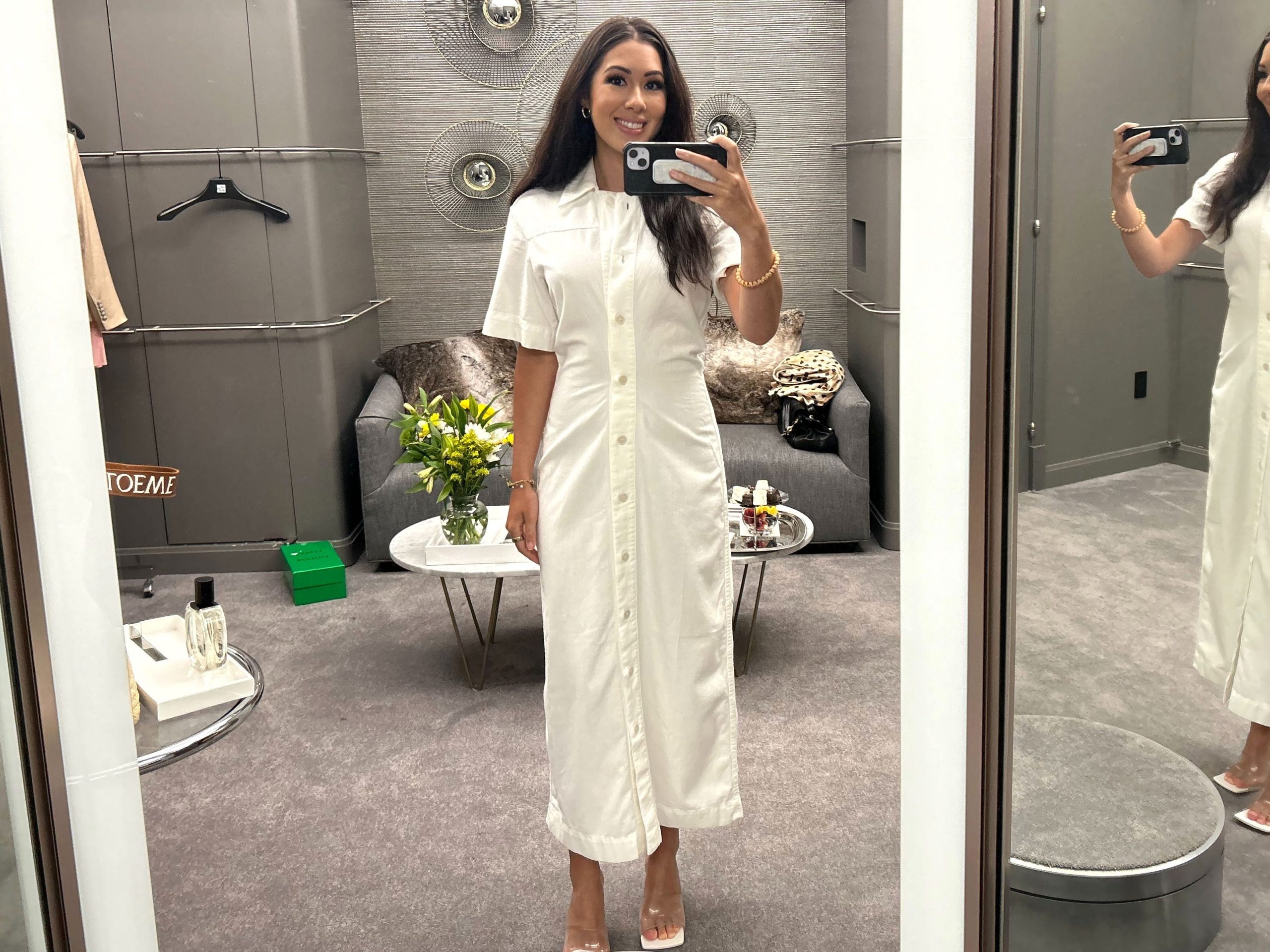 Lara takes a photo of herself in the Saks fitting room wearing a white button-down midi dress with a collar.