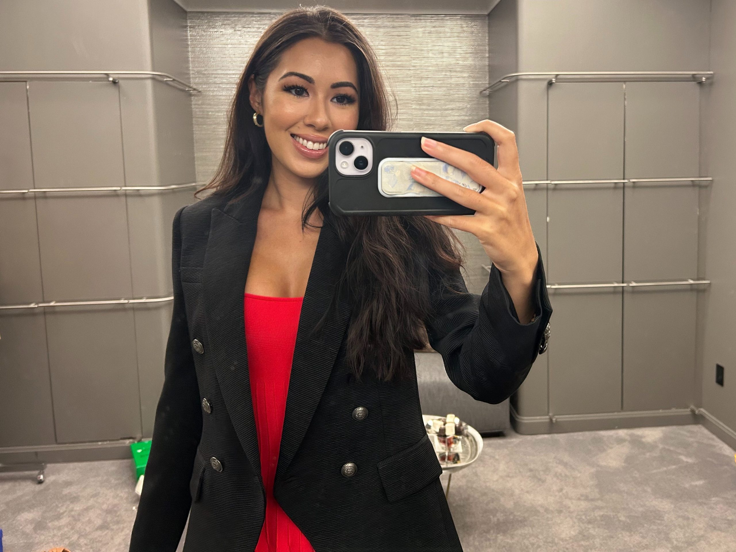 Lara takes a photo of herself wearing a red shirt and a black blazer with buttons.