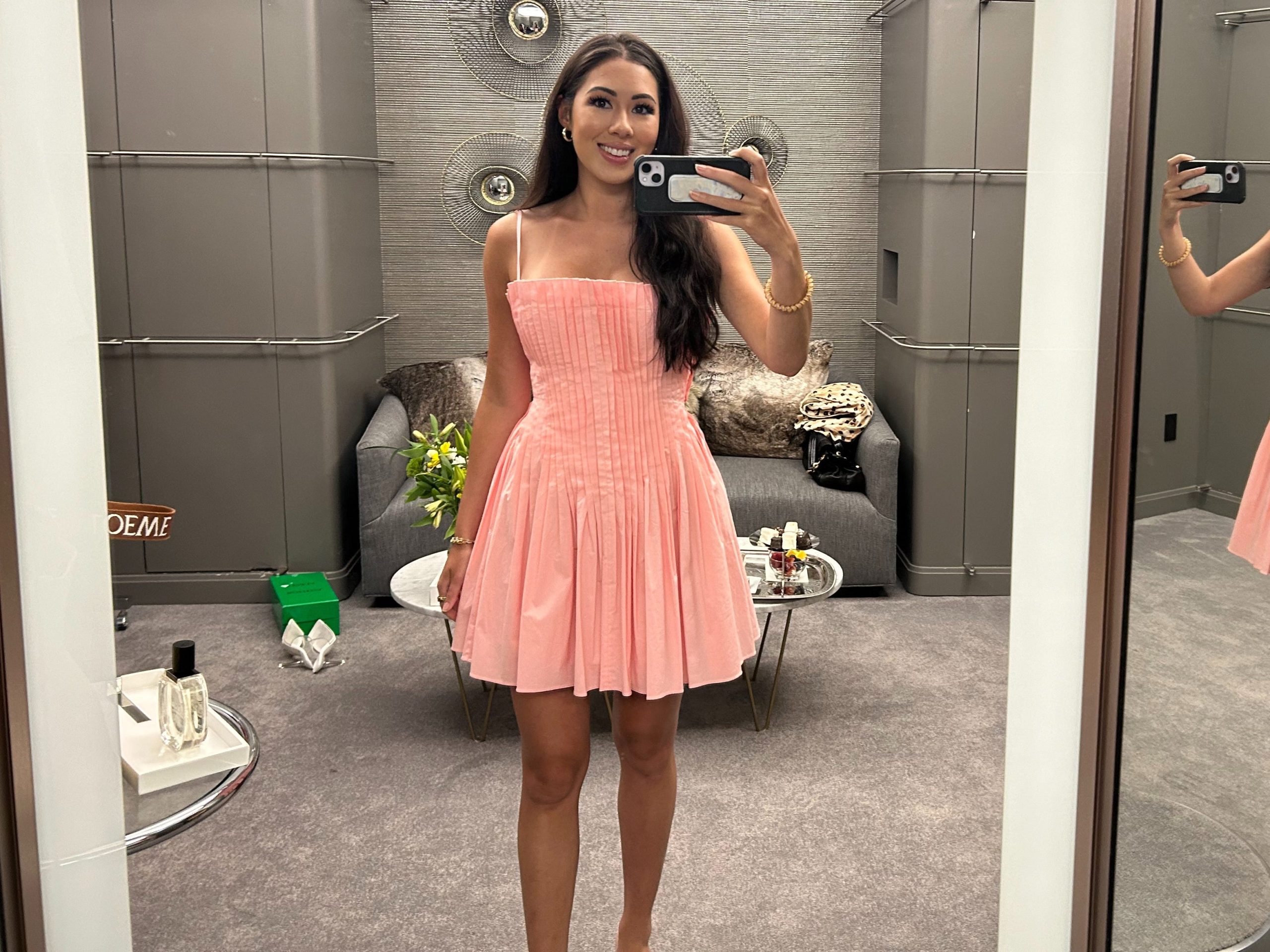 Lara takes a photo of herself in the Saks fitting room wearing a short pink dress.