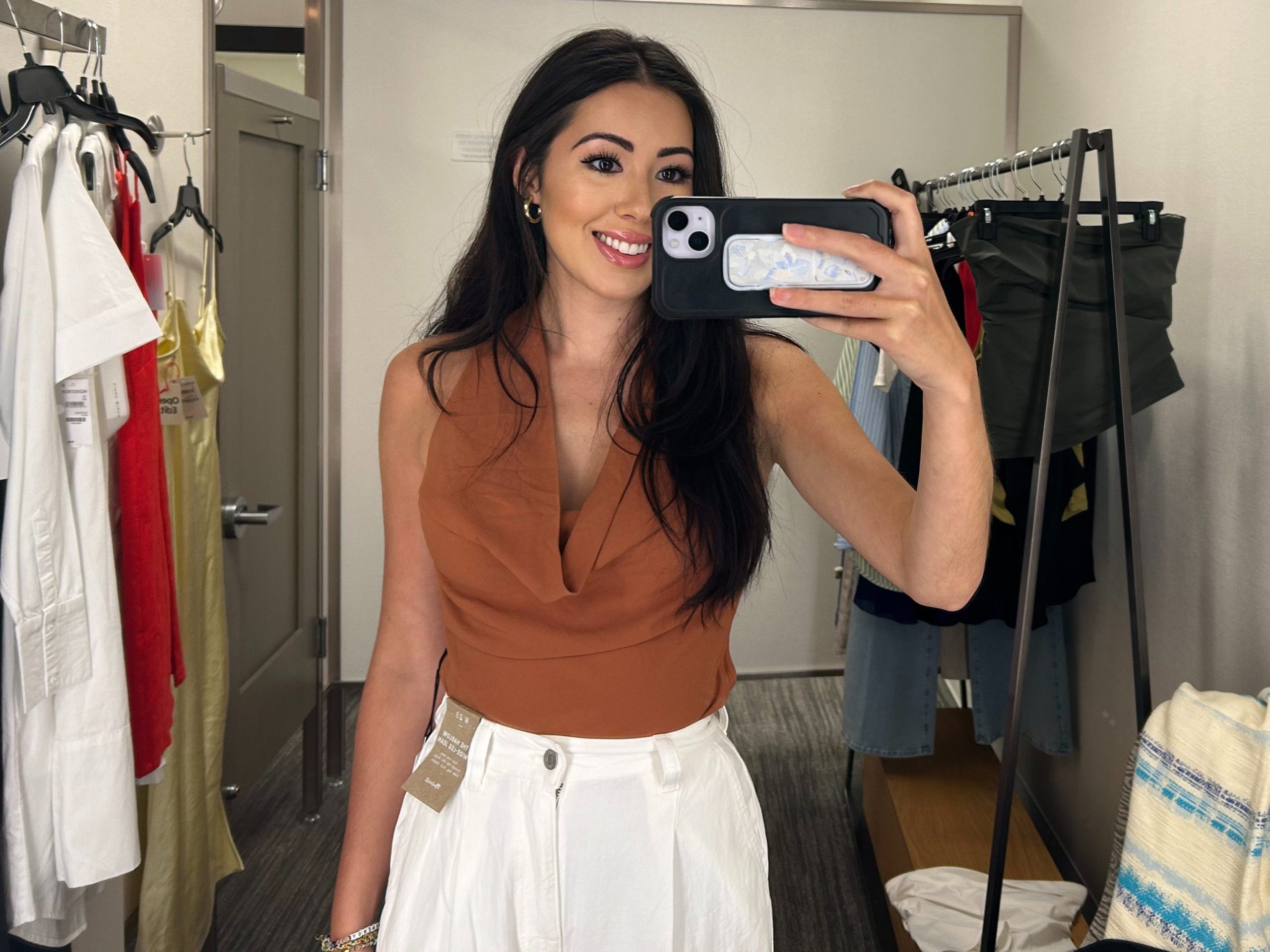 Lara takes a photo of herself in the Nordstrom fitting room wearing a burnt-orange cowl-neck bodysuit and white pants. There are racks of clothing behind her.