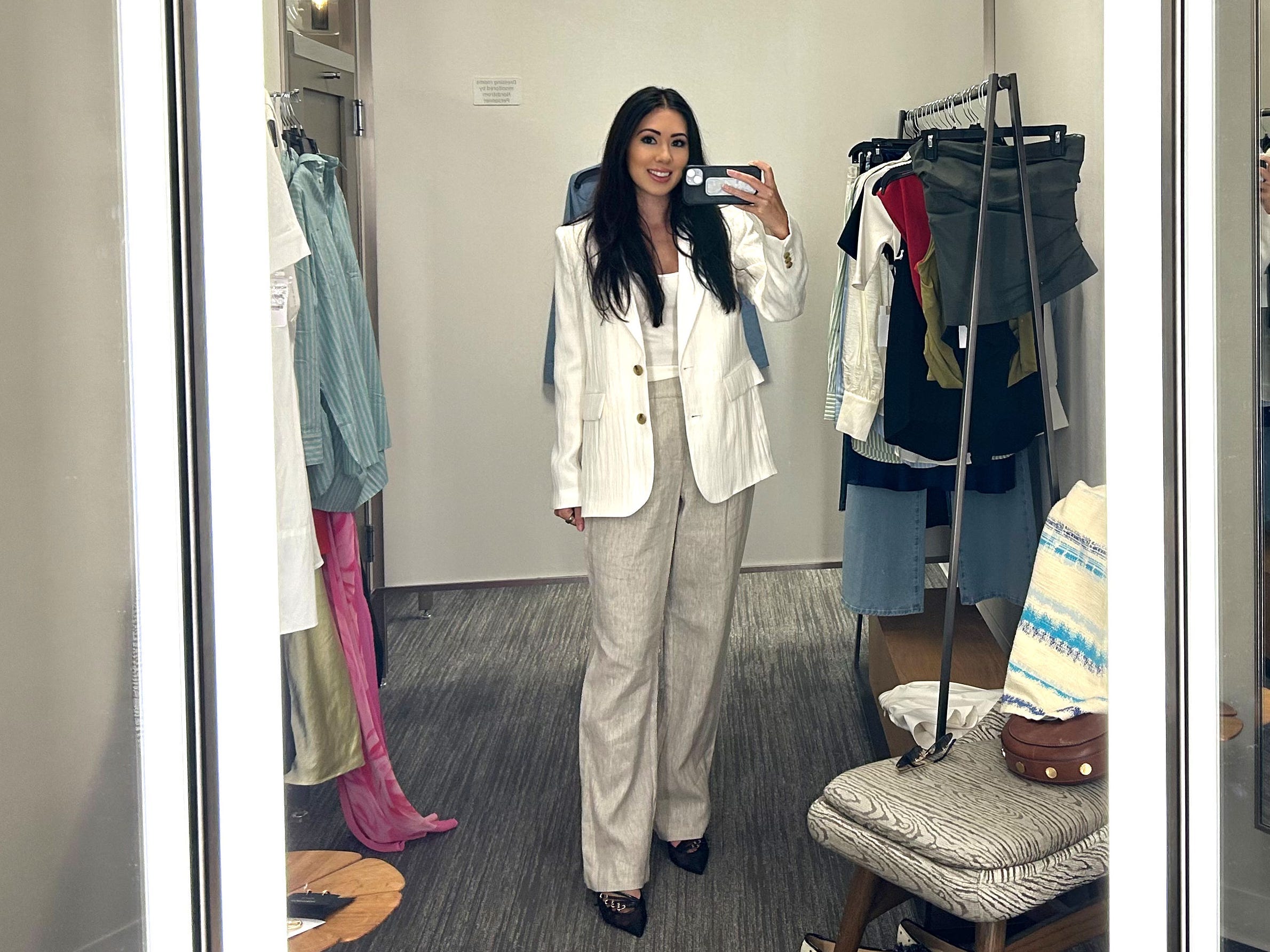 Lara takes a photo in a mirror wearing a cream blazer, white shirt, and gray trousers. There are racks of clothing on the side of her.