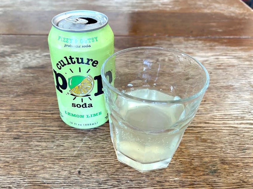 An open can of lemon-lime Culture Pop next to a small, clear glass with light yellow liquid inside. Both are sitting on a wooden table.