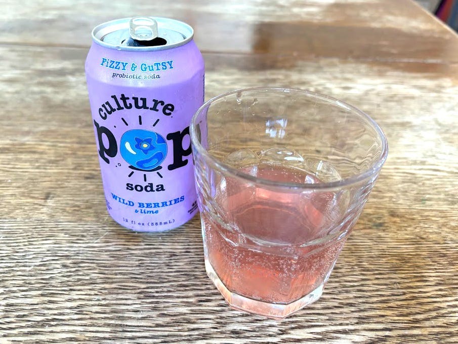 An open can of wild-berries-and-lime Culture Pop next to a small, clear glass with pink liquid inside. Both are sitting on a wooden table.