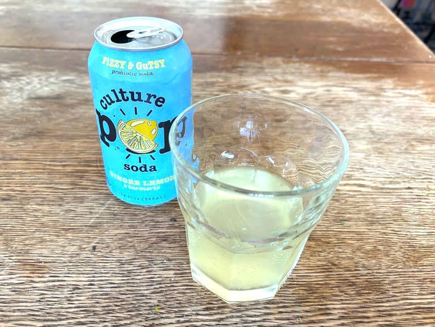 An open can of ginger-lemon-and-turmeric Culture Pop next to a small, clear glass with yellow liquid inside. Both are sitting on a wooden table.